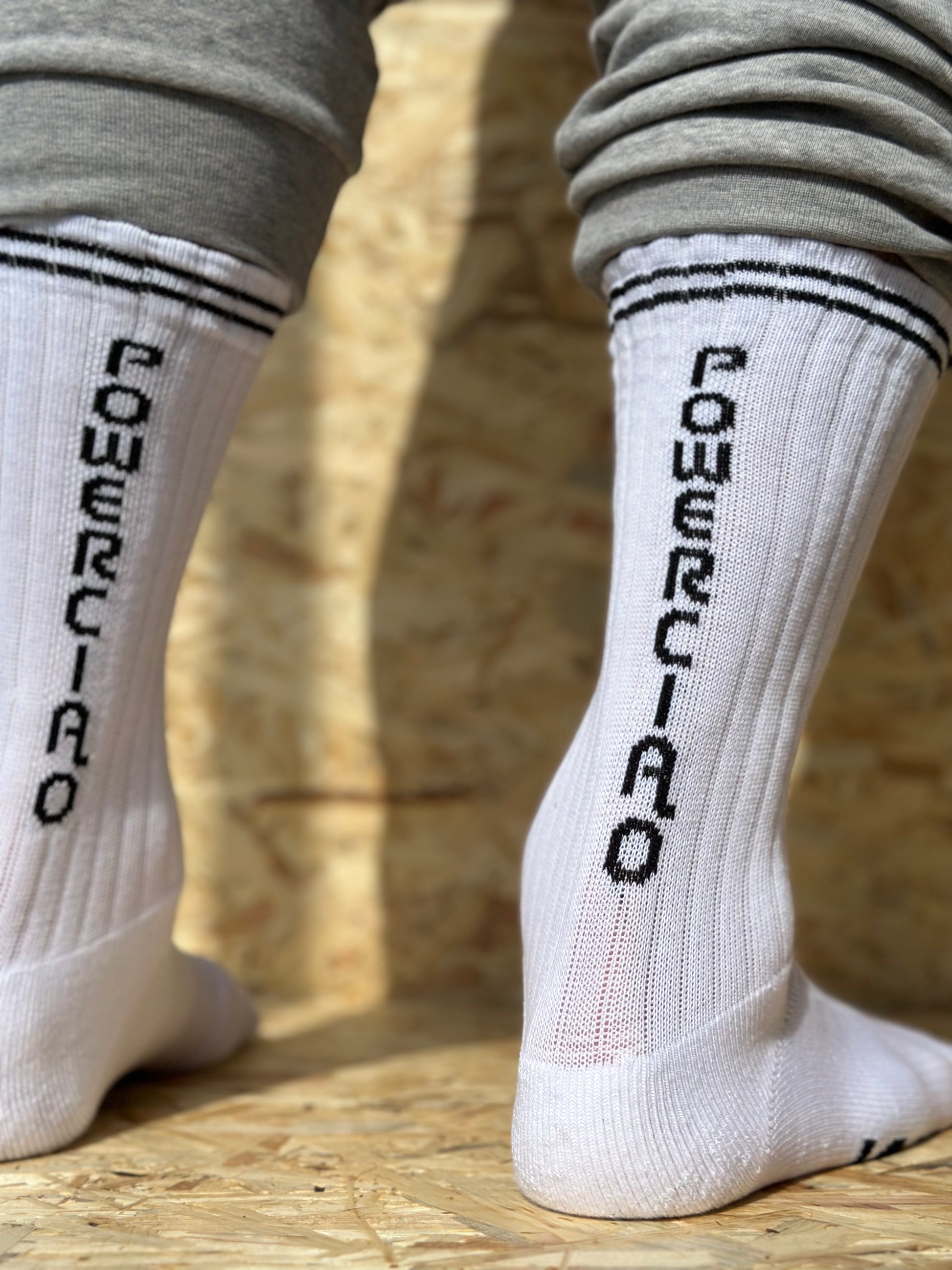 Close-up view of a person's feet wearing white high-calf socks with "Powerciao" written in black vertically on the back, standing on a wooden surface, perfect for strutting around like a sustainable sock superstar.